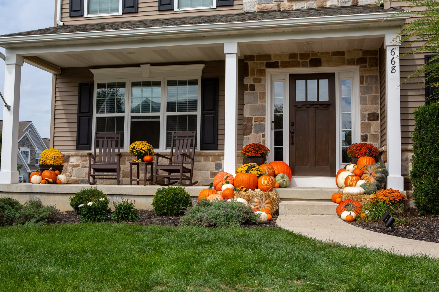 A porch on a stone-front colonial banked with pumpkins in orange, white and green, two straw bales, mums, and custom engraved pumpkins down the steps and along the front walk