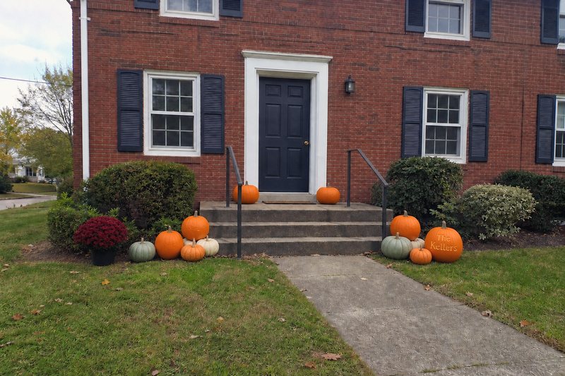 The Signature Porch on a brick colonial entry: an orange pumpkin either side of the door, the rest grouped on the mulch at the foot of the steps with a burgundy mum on the left and one custom engraved pumpkin on the right