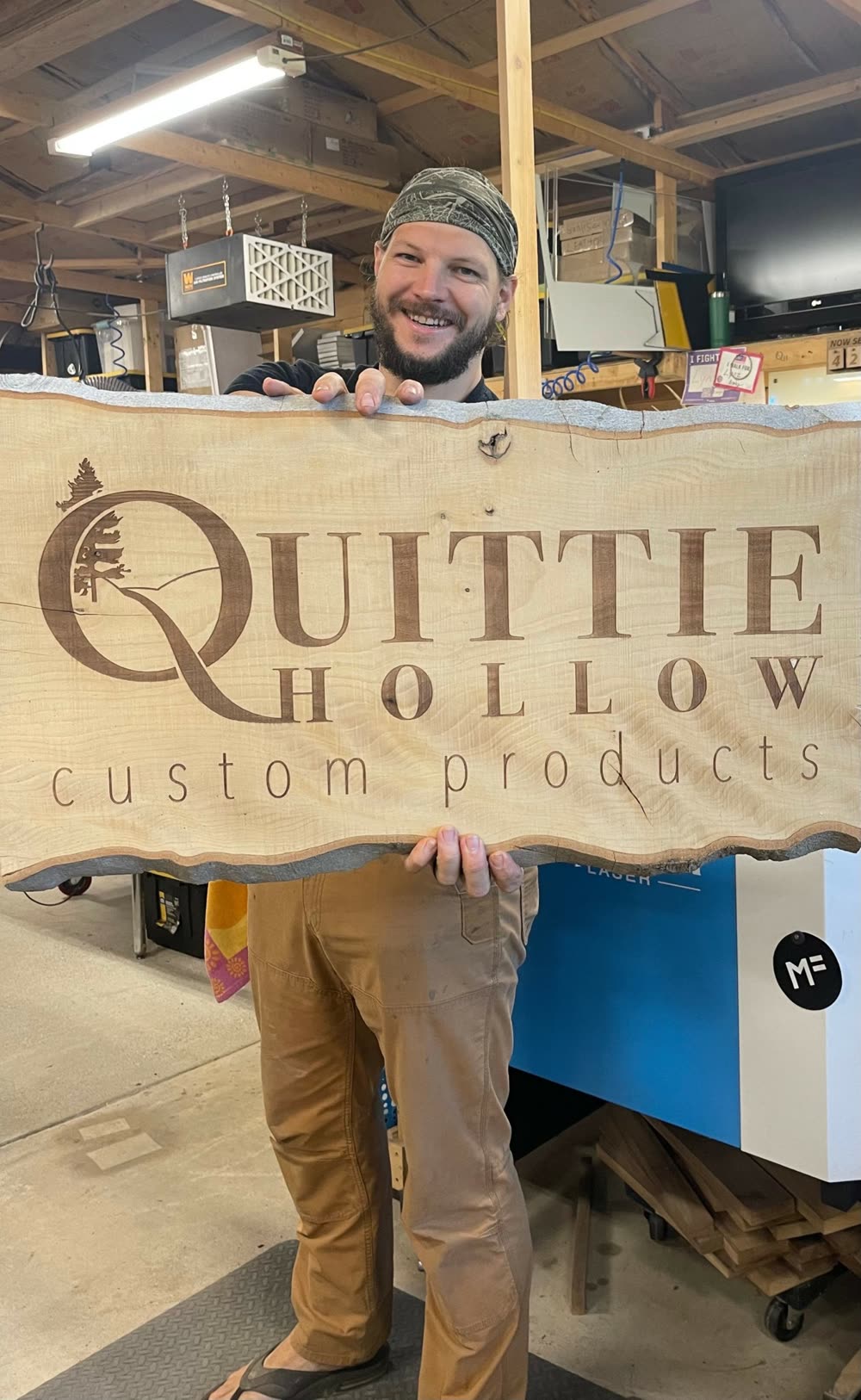 Mike Reese in the Quittie Hollow workshop holding a live-edge slab engraved with the workshop logo, a laser engraver behind him