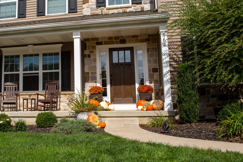 The Signature Porch on a stone-front colonial: a dozen pumpkins in orange, white and green either side of the door around the engraved one, an orange mum each side